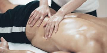 A person is lying face down while receiving an oil massage on their lower back from a massage therapist with both hands. The person is wearing black athletic shorts.
