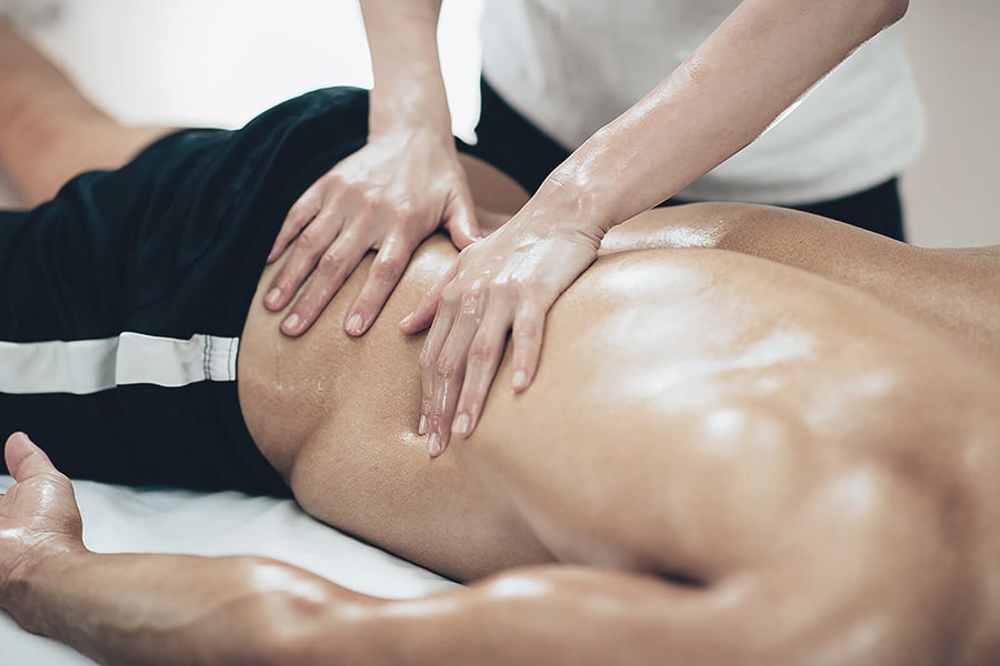 A person is lying face down while receiving an oil massage on their lower back from a massage therapist with both hands. The person is wearing black athletic shorts.